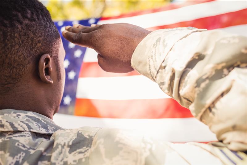 A person in military uniform salutes while facing an American flag outdoors.